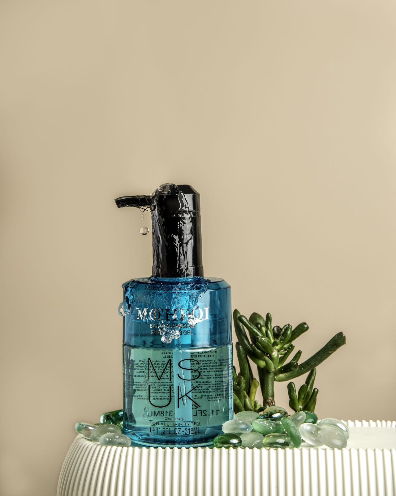 Turkusowa butelka szamponu do włosów MOHEQI MUSK o pojemności 318 ml, postawiona na białym, prążkowanym stole, na beżowym tle. Na szamponie widnieje napis "MUSK" oraz informacja, że przeznaczony jest do każdego rodzaju włosów i zawiera olejek z drzewa herbacianego. Wokół szamponu znajdują się kawałki turkusowego szkła i mały, zielony sukulent.