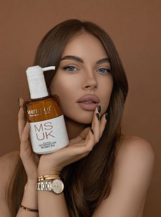 Piękna, młoda kobieta o brązowych, falowanych włosach pozuje, trzymając w dłoni butelkę kosmetyku do włosów. Na butelce z pompką widnieje napis „MUSK” oraz „M@HEQI”. Kobieta patrzy bezpośrednio w obiektyw, z poważnym wyrazem twarzy. Tło jest jednolicie brązowe.